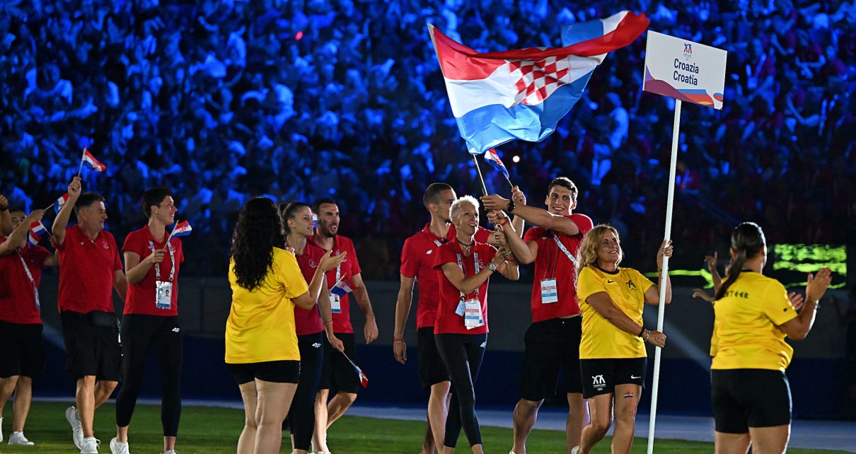 A group of people in red and yellow attire walk across a field, waving a Croatian flag and holding a sign that reads "Croatia," with a large crowd illuminated in blue lights in the background.