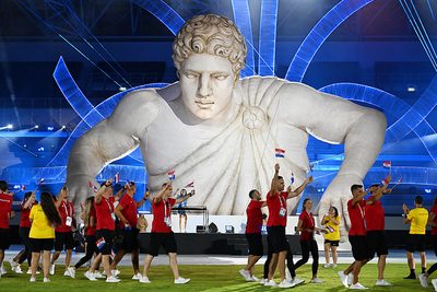 Athletes in red shirts wave flags as they march past a large inflatable statue of a muscular figure on a stage with blue lighting.